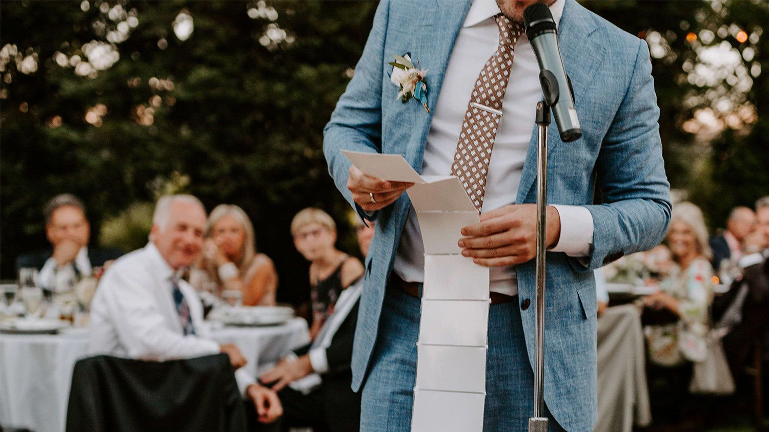 Man in a blue suit standing at a microphone with a blurred background of people seated at tables.